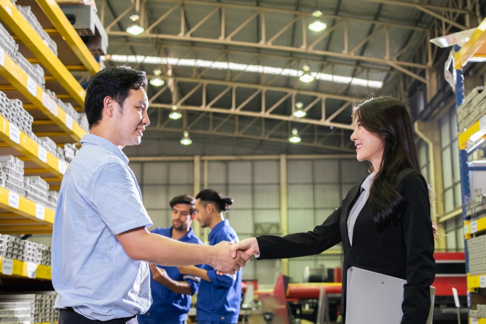 A businesswoman shaking hands with a factory worker inside a warehouse, symbolizing international trade collaboration.
