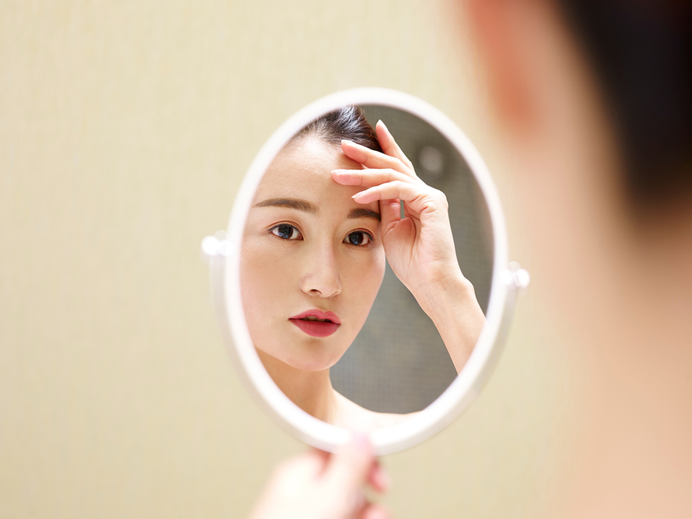 Asian woman examining her face in a hand mirror, symbolizing consumer trends and opportunities in Vietnam’s cosmetics industry